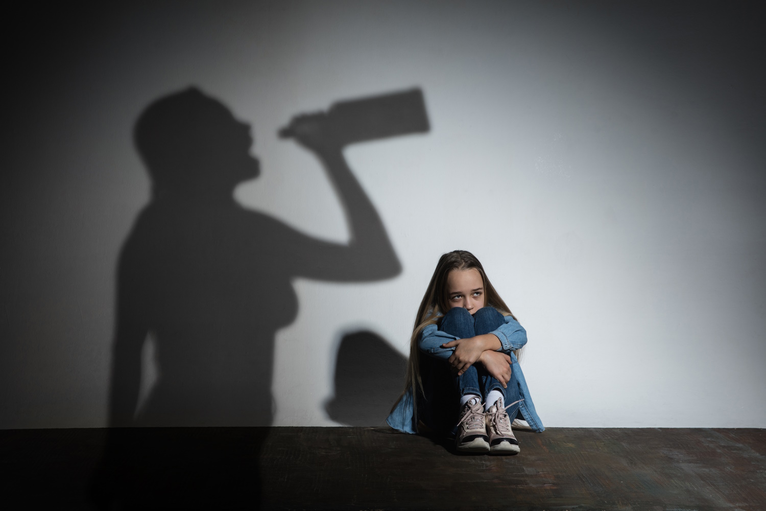 Child sitting sadly against wall with shadow of person drinking cast against the wall